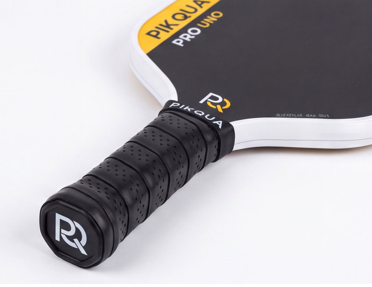 Close-up of a Piqua Pro Uno Kevlar pickleball paddle showing the black perforated handle grip with a white and black bottom cap logo, and part of the black paddle face with yellow and white branding on a clean white background.