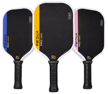 Three pickleball paddles shown side by side on a white background: a blue-bordered Pikaqua Pro Uno Kevlar, a yellow-bordered Pikaqua Pro Uno Kevlar, and a pink-bordered Pikaqua Pro Uno Kevlar. Each paddle has a black face, white edge trim, black wrapped handle, and visible IPMA certification and Pikqua branding.