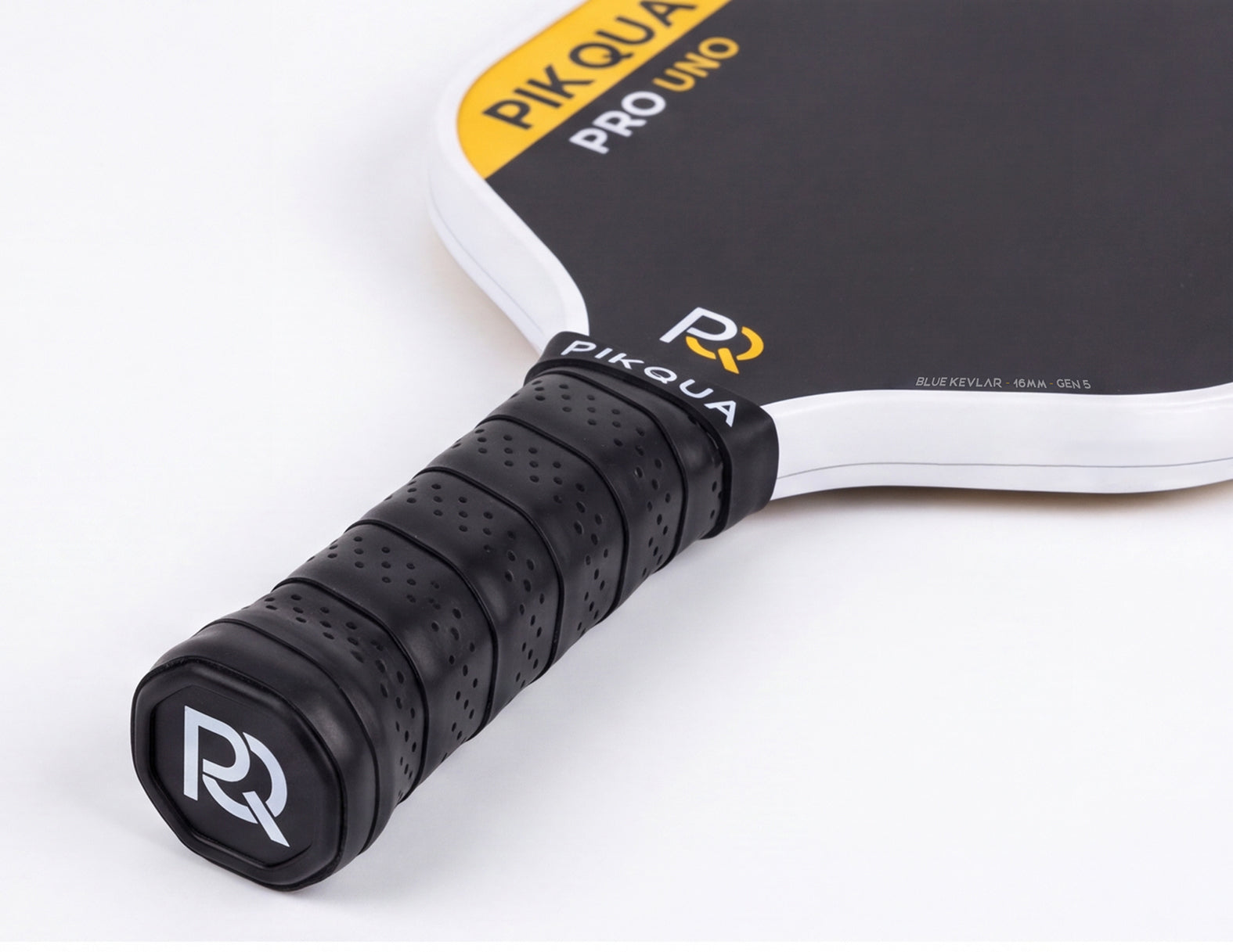 Close-up of a Piqua Pro Uno Kevlar pickleball paddle showing the black perforated handle grip with a white and black bottom cap logo, and part of the black paddle face with yellow and white branding on a clean white background.
