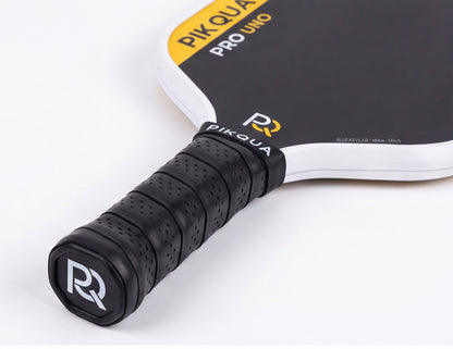 Close-up of a Piqua Pro Uno Kevlar pickleball paddle showing the black perforated handle grip with a white and black bottom cap logo, and part of the black paddle face with yellow and white branding on a clean white background.